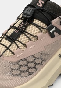 Ελαφρύ παπούτσι για μονοπάτια με υφή κυψελωτού μοτίβου σε μπεζ, ροζ λεπτομέρειες και μαύρα εξαρτήματα· διαθέτει ελαστικά κορδόνια και σόλα με μαξιλαράκι.