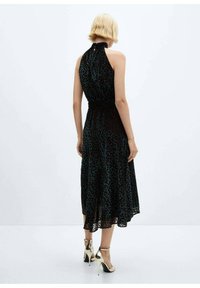 Schwarzes Kleid mit einem Halterneck, gemustert mit türkisfarbenen Pünktchen. Elastischer Taillenbund, schwingender Rock und durchscheinende Textur. Tragen mit beigen Absätzen.