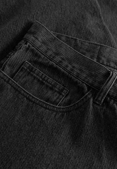 Fekete farmernadrág, texturált anyaggal, látható hátsó zsebbel és erős derékpánttal, amelyhez övbújtatók vannak.