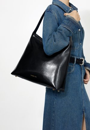 Osoba u plavom traper kaputu i crnom remenu, nosi veliku crnu kožnu torbu preko ramena sa zlatnim nogicama i suptilnim natpisom brenda.