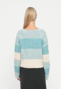 Žena s dlouhými blond vlasy, oblečená v pleteném svetr s pastelovými pruhy v světle modré, krémové a šedé barvě, v kombinaci s tmavou sukní, viděná zepředu.