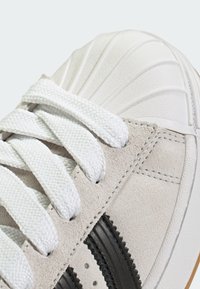 Biele tenisky s textúrovanou špičkou, semišovým zvrškom v svetlosivej farbe, bielymi šnúrkami a čiernymi pruhmi. Viditeľná gumová podrážka a švy.