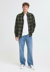 Camicia flanella a quadri verdi e neri sopra una maglietta bianca, abbinata a jeans a gamba larga blu chiaro e sneakers bianche con strisce nere.