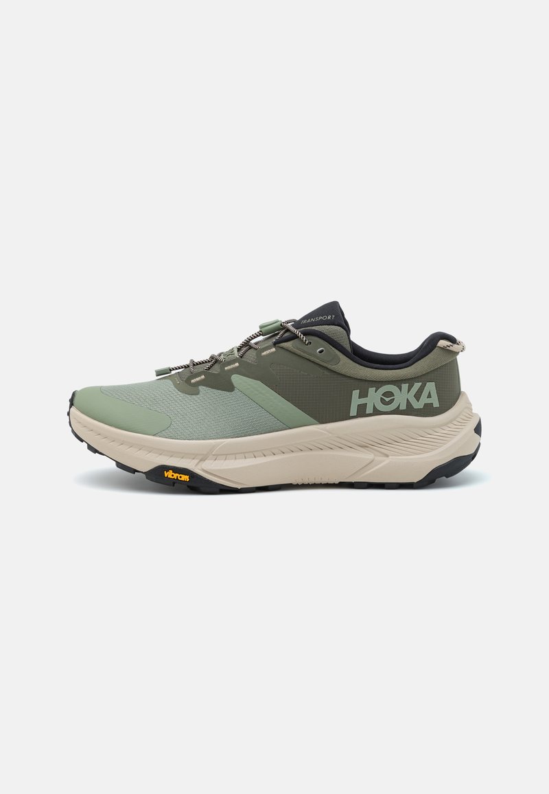 Zapatilla de correr Hoka con parte superior de malla verde, entresuela Beig, acentos negros y suela Vibram. Presenta un sistema de lazado único.