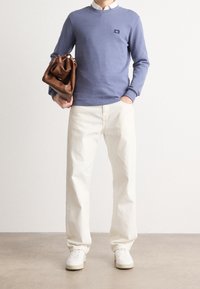 Person som bär en ljusblå tröja, vit skjorta med krage, vita byxor, vita sneakers, håller en brun läderkuvertväska mot en neutral bakgrund.