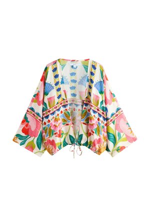 Vrouw met lang donker haar, gouden oorbellen, een kleurrijk bloemenkimono over een wit tanktopje en witte broek, staand tegen een gestructureerde muur.