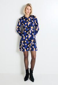 Vestido de manga larga en patrón geométrico azul y beige; escote con cremallera, de largo por encima de la rodilla, hecho de tejido texturizado. Botines negros.