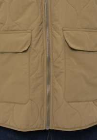 Μπουφάν σε μπεζ χρώμα με καπιτονέ σχέδιο, διαθέτει φερμουάρ μπροστά, δύο πλαϊνές τσέπες και υφασμάτινα μοτίβα με υφή. Το υλικό είναι απαλό και ελαφρώς γυαλιστερό.