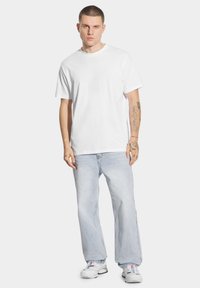 Biała bawełniana koszulka, luźny krój, krótkie rękawy, zestawiona z jasnoniebieskimi dżinsami, prostymi nogawkami, noszona z białymi sneakersami, widoczne tatuaże na ramieniu.