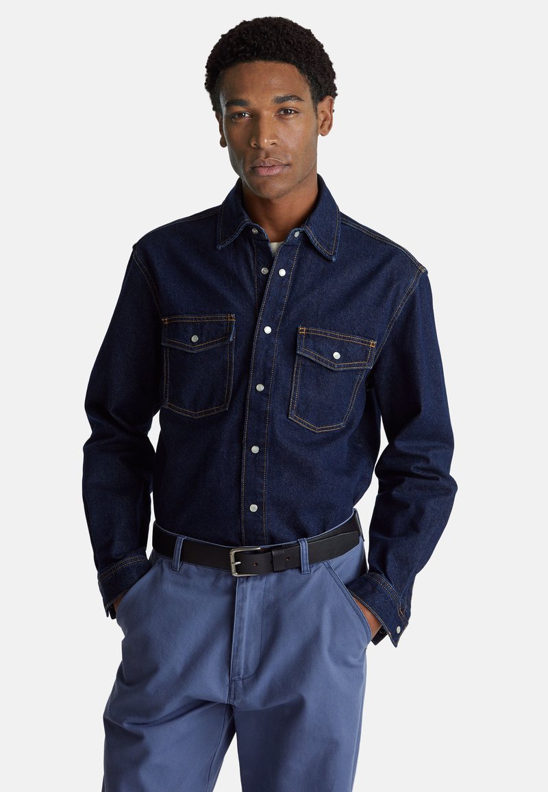 Camicia in denim scuro con due tasche sul petto, chiusura a bottoni e cuciture chiare. Abbinata a pantaloni larghi di un blu chiaro e una cintura nera.