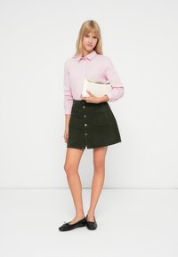 Nainen pukeutuneena vaaleanpunaiseen raidalliseen paitaan, tummansiniseen napitettuun hameeseen ja mustiin balleriinoihin, pitää valkoista clutch-laukkua, seisoo valkoista taustaa vasten.