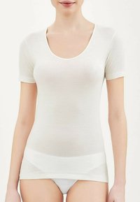 Top bianco a maniche corte realizzato in tessuto leggero e trasparente, con scollatura a barca e texture liscia, indossato sopra biancheria intima bianca.