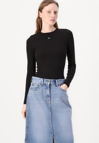 Svart, langermet tettsittende topp kombinert med en lys blå denimskjørt med en frontsliss og frontlommer, akkompagnert av en liten logo.