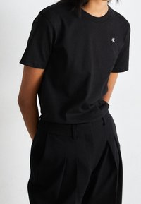 Personer klädd i en svart kortärmad t-shirt med en liten vit "CK"-logotyp på bröstet och svarta veckade byxor, med händerna i fickorna.