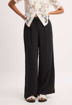 Kobieta ubrana w biały, krótki sweter z dzianiny, czarne szerokie spodnie z troczkiem oraz białe sandały na płaskiej podeszwie, stojąca z rękami na biodrach.