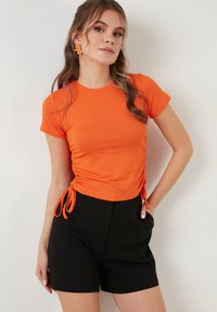 Oranje geribde top met korte mouwen en zijkanten met ruching en strikjes, gecombineerd met zwarte high-waisted shorts. Gladde textuur en aansluitend ontwerp.