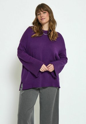 Pull en maille violette avec des poignets et un ourlet côtelés, des fentes latérales et une coupe décontractée, assorti à un pantalon gris ample.