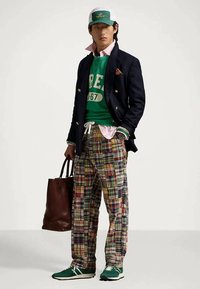 Άνδρας με πράσινο καπέλο, πράσινη ζακέτα με λευκή εκτύπωση, ναυτική μπλέ σακάκι, πολυχρωμο καρό παντελόνι, πράσινα αθλητικά παπούτσια, και κρατώντας μια καφέ δερμάτινη τσάντα.