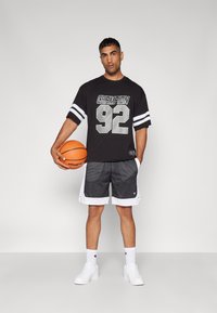 Tricou negru cu mânecă scurtă, având inscripția "CHAMPION 92" în argintiu, combinat cu pantaloni scurți de baschet din plasă gri și adidași albi, ținând o minge de baschet.