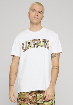 Ung mann med kort blondt hår iført hvit t-skjorte med kamuflasjemønster og teksten "UNFAIR" samt bukser med kamuflasjemønster, står foran en ensfarget bakgrunn.