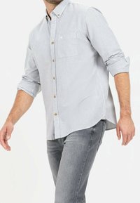 Chemise à rayures bleues et blanches, boutonnée, avec une poche poitrine, manches retroussées et boutons marron, associée à un jean gris.