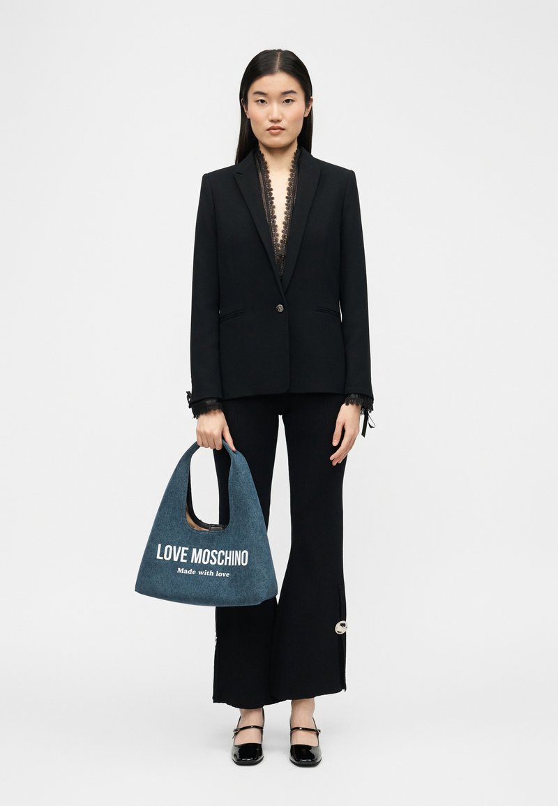 Γυναίκα που φοράει μαύρο σακάκι και παντελόνι, κρατώντας μια μπλε τσάντα Love Moschino από τζιν με λευκό κείμενο, στέκεται μπροστά σε λευκό απλό φόντο.