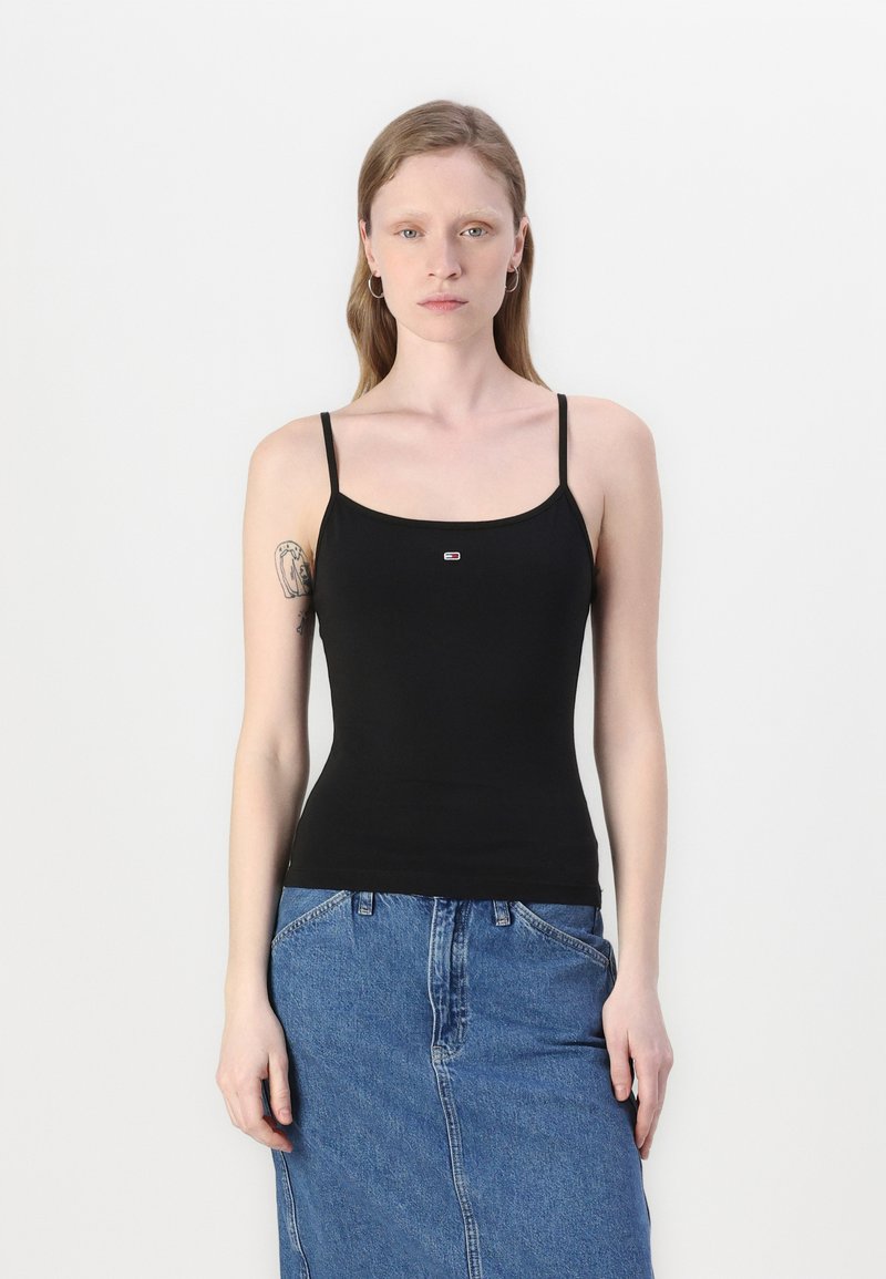 Black fitted tank top with thin straps and small logo detail at the center. Paired with a denim skirt, showcasing a casual ensemble.