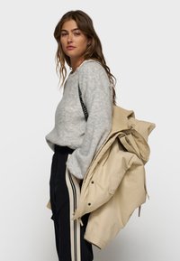 Grå, struktureret sweater med rund hals, iført med mørke bukser med lyse sidelister og en beige jakke draperet over skulderen.