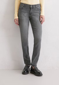 Personne portant un jean gris slim, des chaussures noires à enfiler et un haut à manches longues côtelé jaune clair, se tenant contre un mur blanc texturé.