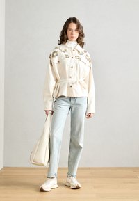 Giacca over-size color crema con ricami floreali, bottoni e cintura, abbinata a jeans azzurri chiari e sneakers bianche. Borsa beige.