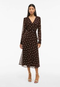 Bruine jurk met polka dots, met lange doorschijnende mouwen, een V-halslijn en strikdetail in de taille, met een zwierige midi-lengte en lichtgewicht stof.