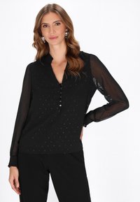 Blusa nera con maniche lunghe trasparenti, patta con bottoni, tessuto strutturato caratterizzato da piccoli motivi e colletto a punta. Indossata con pantaloni neri.