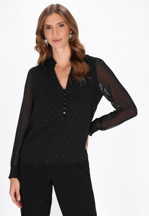 Schwarze Bluse mit langen durchsichtigen Ärmeln, Knopfleiste, strukturiertem Stoff mit kleinen Mustern und einem spitzen Kragen. Gefeit mit schwarzen Hosen.