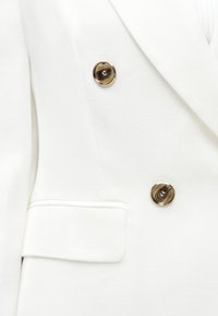 Λευκό σακάκι διπλής σειράς με καφέ κουμπιά με χρυσό περίγραμμα και τσέπη με καπάκι στην αριστερή πλευρά.