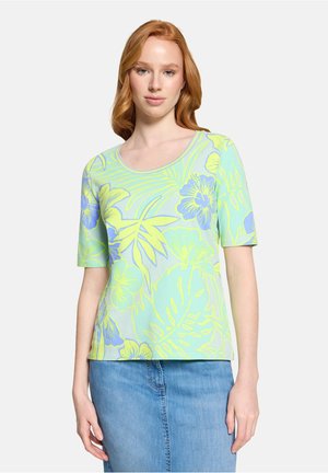 Femme aux cheveux roux portant un haut à manches courtes avec des fleurs pastel vertes et bleues, et une jupe en jean bleue, debout devant un fond uni.