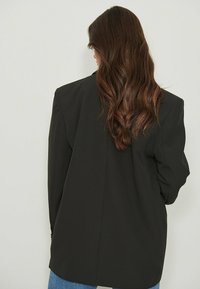Blazer noir avec épaules structurées et coupe ample, présentant une texture lisse et une fente unique au dos.