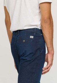 Pantalon en denim bleu foncé avec une finition texturée, doté d'une poche arrière avec une étiquette de marque et d'une fermeture à un seul bouton.