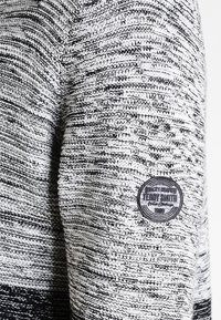 Sweter dziany z fakturowanym, monochromatycznym wzorem w czerni i bieli. Na rękawie znajduje się okrągła naszywka z logo i szczegółami tekstowymi.