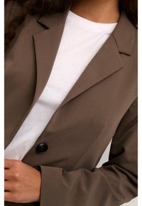 Ruskea räätälöity bleiseri, jossa on yksi musta nappi, lovettu kaulus ja sileä kangas, päällä valkoinen pyöreäkauluksinen paita.