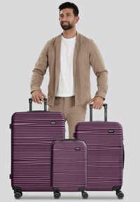 Un homme en tenue beige se tient derrière trois valises rigides violettes de différentes tailles avec des poignées extensibles sur un fond gris.