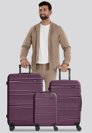 Hombre vestido con un atuendo beige se encuentra detrás de tres maletas rígidas moradas de diferentes tamaños, con mangos extendidos, sobre un fondo gris.