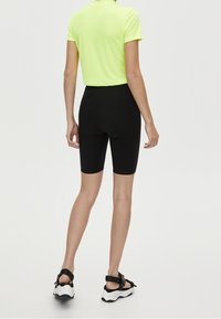 Osoba nosząca neonowożółtą koszulkę z krótkim rękawem, czarne przylegające szorty rowerowe oraz czarno-białe sportowe sandały, stojąca na tle jednolitym.