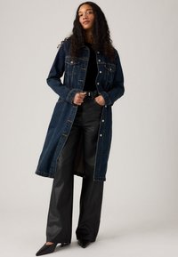 Manteau en denim bleu foncé, doté d'une fermeture à boutons sur le devant, de deux poches poitrine et d'une coupe droite. Porté sur un pantalon noir ajusté et des escarpins pointus.