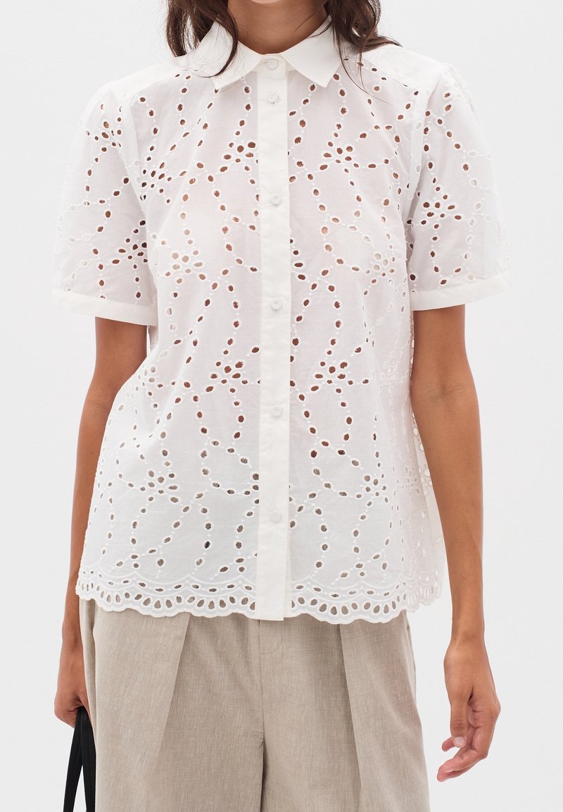 Blouse blanche à manches courtes en dentelle à œillets avec fermeture boutonnée sur le devant, ourlet festonné, portée avec un pantalon plissé beige et un sac à main noir.