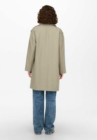 Beige, knielanger Mantel mit strukturiertem Kragen, langen Ärmeln und lockerer Passform, kombiniert mit hellblauen, weit geschnittenen Jeans. Glatte Textur.