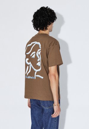 Maglietta di cotone marrone con disegno stilizzato di un volto in linea blu sul retro, abbinata a jeans in denim blu.