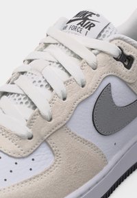 Gros plan sur une sneaker Nike Air Force 1 blanche et beige montrant les lacets, la languette en mesh, les panneaux en daim et cuir, ainsi que le logo Nike swoosh gris.