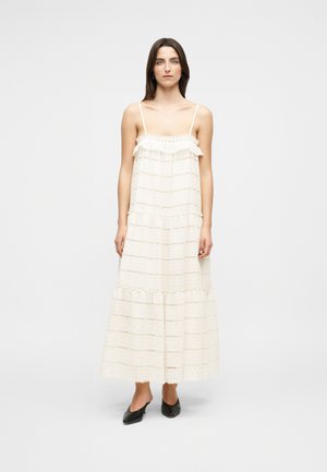 Frau mit langen dunklen Haaren, die ein ärmelloses, gestuftes weißes Maxikleid mit dünnen Trägern und schwarze spitze High Heels trägt und vor einem weißen Hintergrund steht.