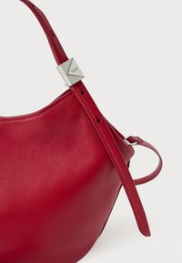 Czerwona skórzana torba o gładkiej fakturze, zaokrąglonym kształcie i srebrzystych elementach ozdobnych. Posiada regulowany pasek z otworami.