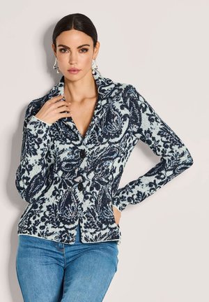 Frau in einem blau-weißen, gemusterten Knopfjacke mit Kragen und blauen Jeans, die mit einer Hand auf der Hüfte und der anderen Hand am Hals posiert.